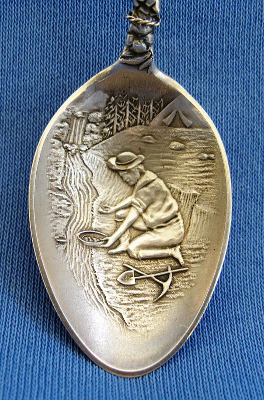 Nevada City CA Gold Panning Bowl.JPG - SOUVENIR MINING SPOON NEVADA CITY CA - Sterling demitasse spoon with embossed bowl showing miner panning a stream with a crossed shovel and pick in foreground, top of handle shows a bear looking over a gold pan with nuggets, marked CALIFORNIA around edge of pan and NEVADA CITY in bottom of pan, crossed pick and shovel behind pan with silver rope wrapped around decorative handle, reverse marked Sterling, 4 3/8 in. long  [Located in California's Sierra Nevada, Nevada City is the county seat of Nevada County, 60 miles northeast of Sacramento.  Originally a mining camp founded along Deer Creek in 1849, Nevada City was known by many monikers through its history including Caldwell's Upper Store, Coyoteville, and Deer Creek Dry Diggings. The name Nevada was adopted in May 1850 at a public meeting, having derived its name after the Spanish word for snow. City was added later after Nevada became a state.  After gold was discovered in Deer Creek, Nevada City rapidly became the largest and wealthiest mining town in California, with 10,000 residents and the third largest city in California at the time.  During the period of 1848-1965, Nevada County produced the largest amount of gold in California at over $440 million.  During the gold rush period, Nevada City was known for its coyote or drift diggings, while its sister town Grass Valley became the center for lode mining.  Nevada City maintained a long history of gold mining operations with 16 major mines in the area.  Mining in the area also changed the landscape dramatically.   Hydraulic mining, where miners aimed high-pressure hydraulic monitors, like large water cannons, at the hillsides to strip away gravel in search of gold, was first practiced in California at American Hill here in 1852 by E. G. Matteson before environmental concerns stopped the practice in the 1880s.]